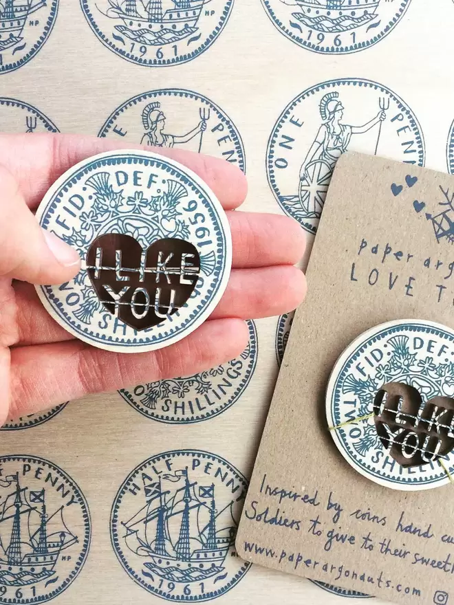 A hand holds the intricate wooden screenprinted Love token coin. The cut out words read 'I like you'