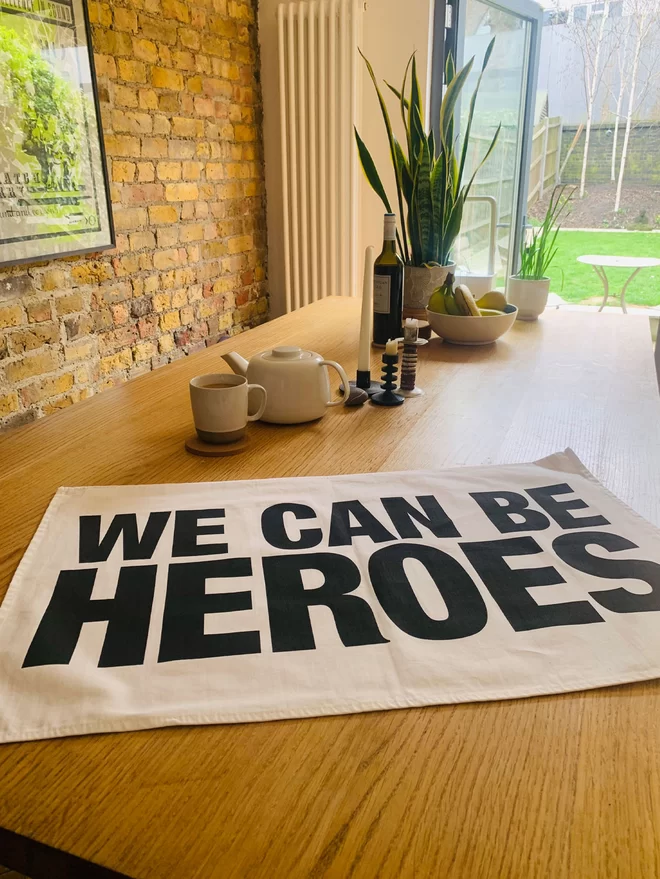 White tea towel with black print saying We Can Be Heroes laying on a timber kitchen island with tea pot, fruit bowl, plant in background