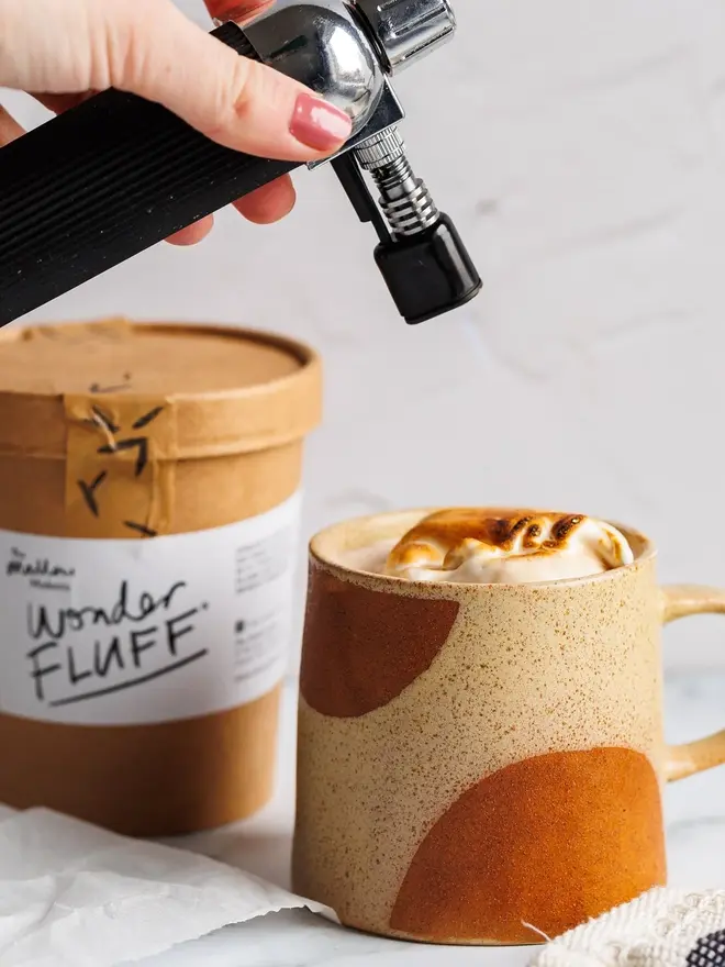 Vanilla Wonder Fluff Tub, a hot chocolate with fluff being cooked on top. This is on a white surface with the tub in the background. 