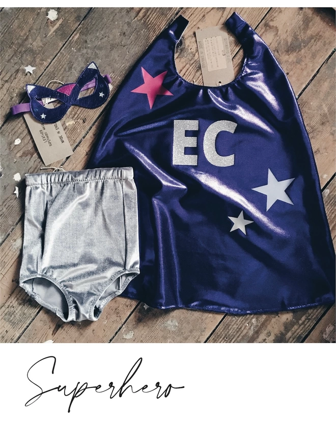 Superhero Costume lying flat on the floor, Purple cape with silver initials and star design, silver grey shorts and purple leather cat eye mask