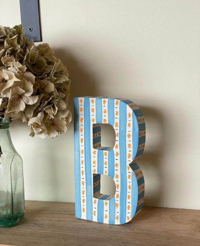 A hand-painted decorative initial letter  with stripes on a wooden table against a light wall. 