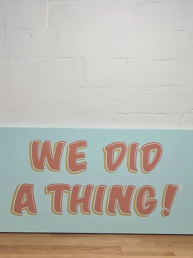'We Did A Thing' Colourful Wedding Sign, a colourful wedding sign sitting on a wooden surface against a white brick backdrop. 