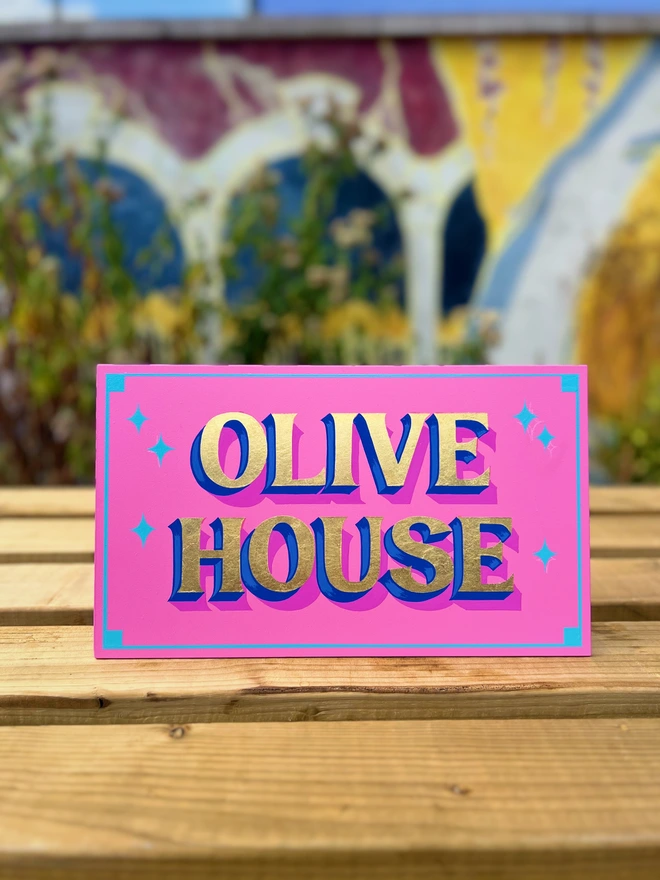 Pink sign with gold leaf lettering spelling 'Olive House', with a deep purple shade and blue stars.