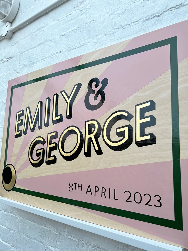 Modern wedding sign featuring the couple's names in gold leaf, outlined in black, and the wedding date, with a sunburst background and a border. Shown against a white brick wall, at an angle. 