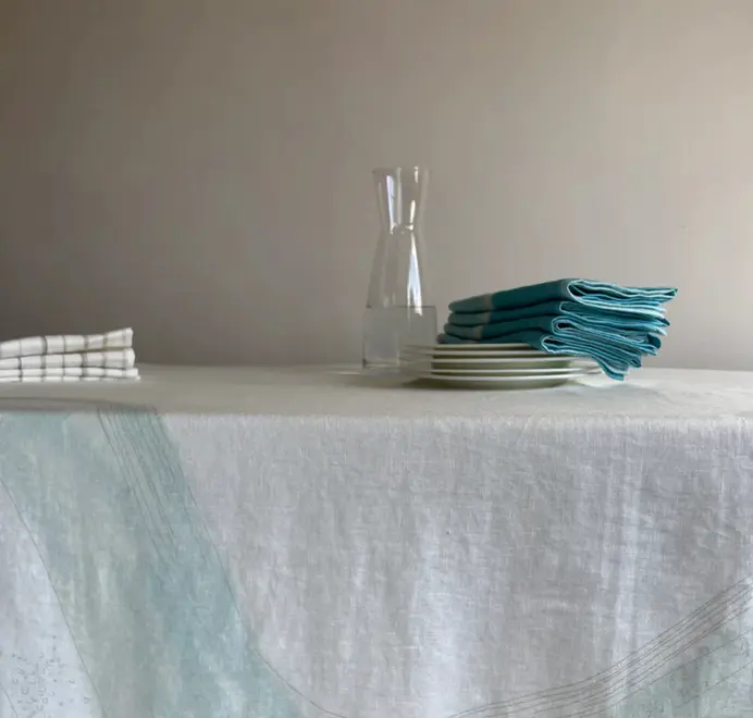 Imola Tablecloth. Light blue and white tablecloth with subtle abstract patterns, styled on an outdoor dining table with plates, glassware, and white floral arrangements.