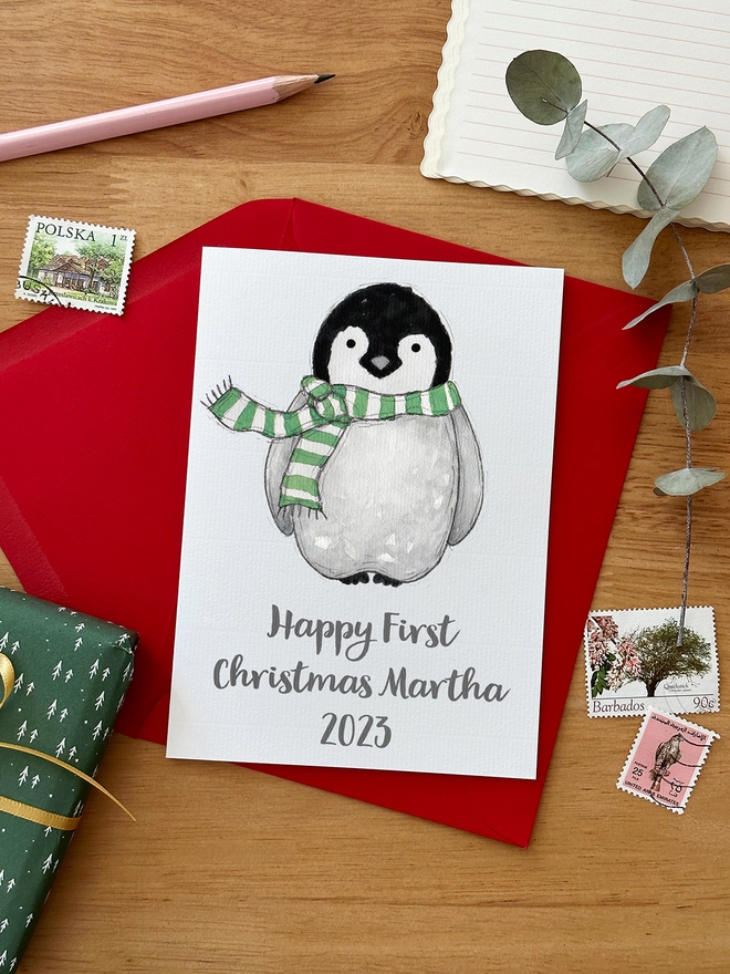 A first Christmas greetings card with an illustrated penguin design lays on a red envelope beside various stationery items on a wooden desk.