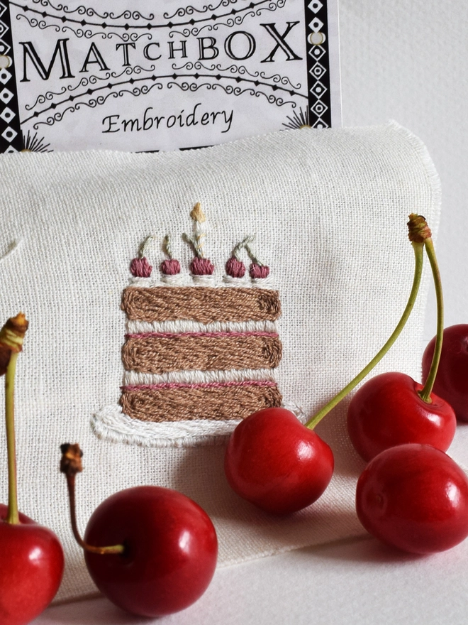 Mini embroidery of a chocolate Black Forest gateau, topped with cherries and a single candle.  Displayed with fresh home grown cherries and the matchbox packaging in the background.