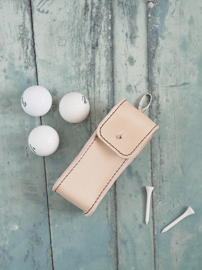 Handmade leather golf ball carry case. A natural cream leather pouch surrounded by 3 golf balls and 2 tees.