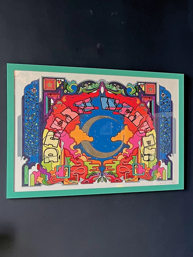 Framed in a teal, hung on a dark grey wall, the image a silver waxing crescent moon sits in the centre under the words “Dream Weaver” which arches around it. The central illustration is brightly coloured, and is edged with a dark blue border.