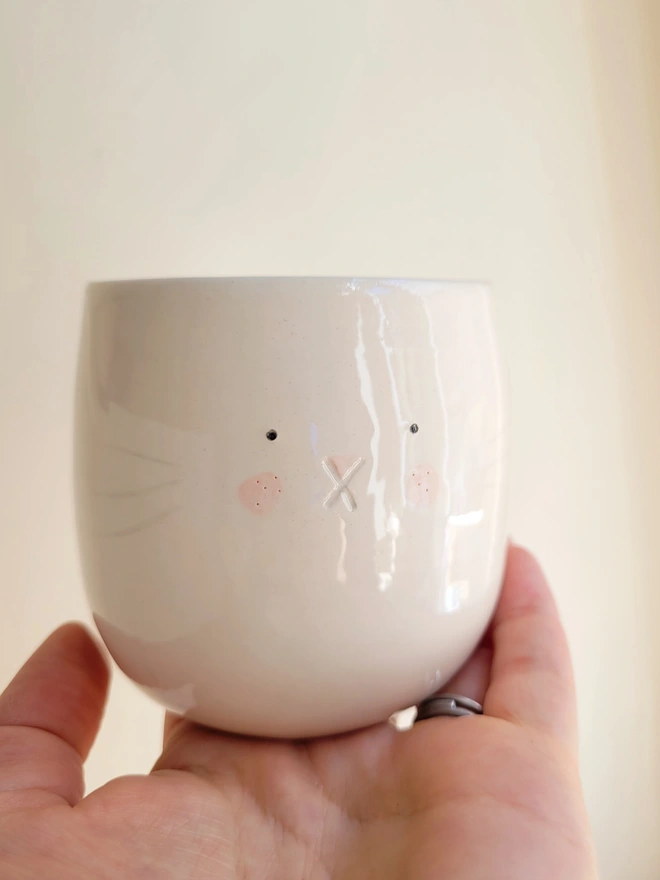 a handmade pottery beaker cup with a cute bunny face being held in a hand