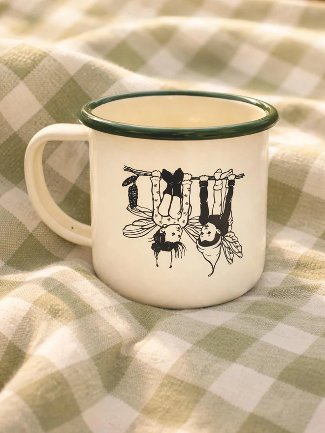 an enamel mug cream with a green rim featuring two pixie pals hanging upside down from a twig sat on a picnic blanket 