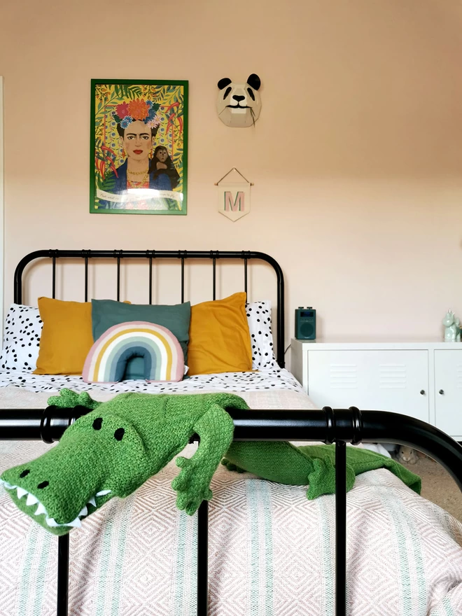 Claude the crocodile long hot water bottle draped over the end rail of a black metal framed double bed, showing his length, drape and scale.