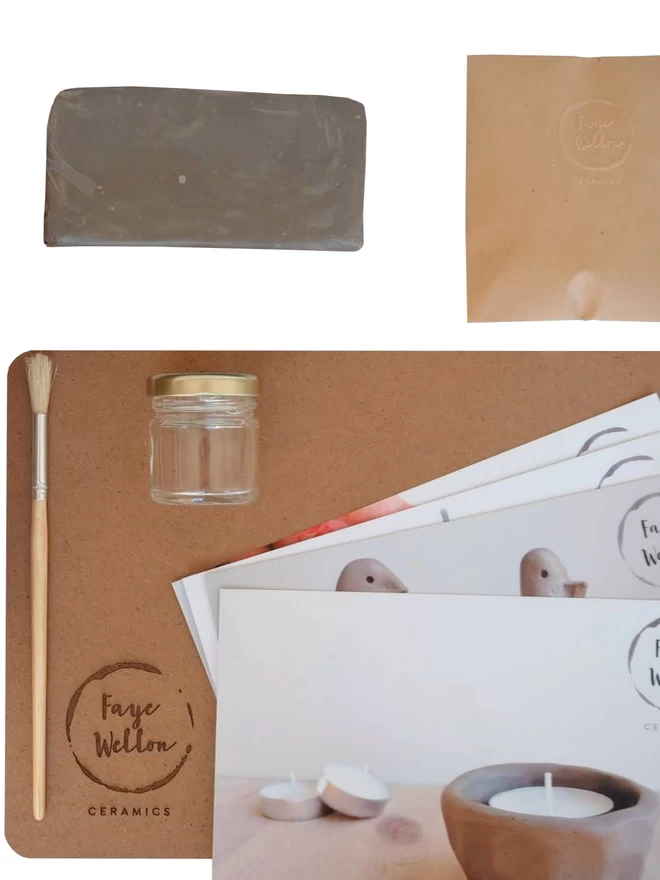 Contents of pottery kit: clay board, instruction cards, paintbrush, jar, clay and envelope. White background