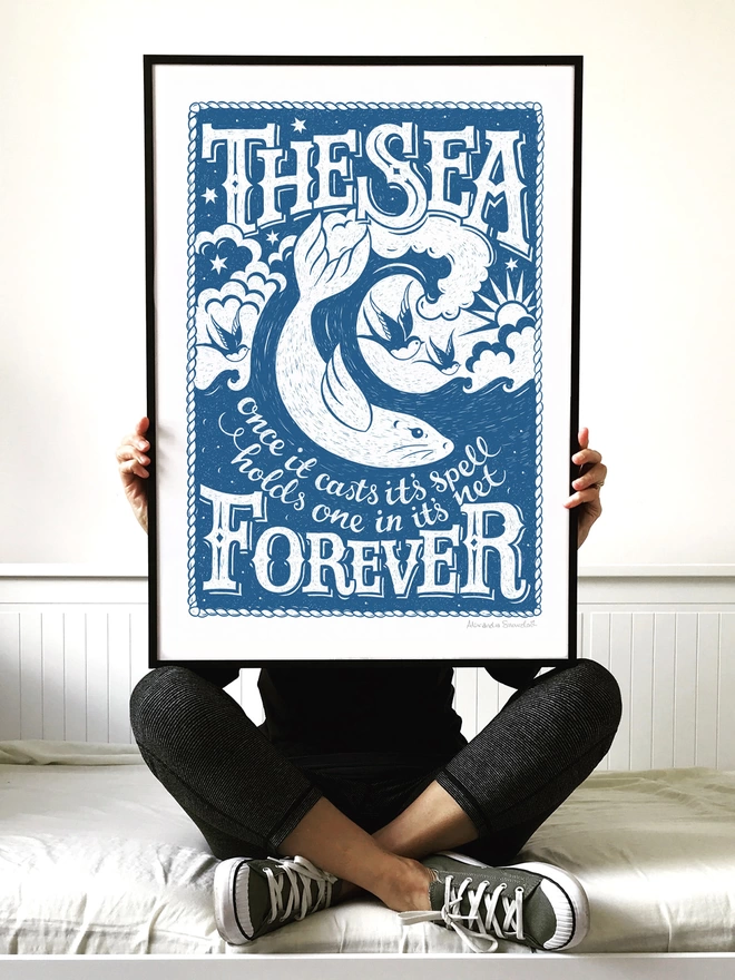 woman sat crossed legged holding blue sea print in black frame