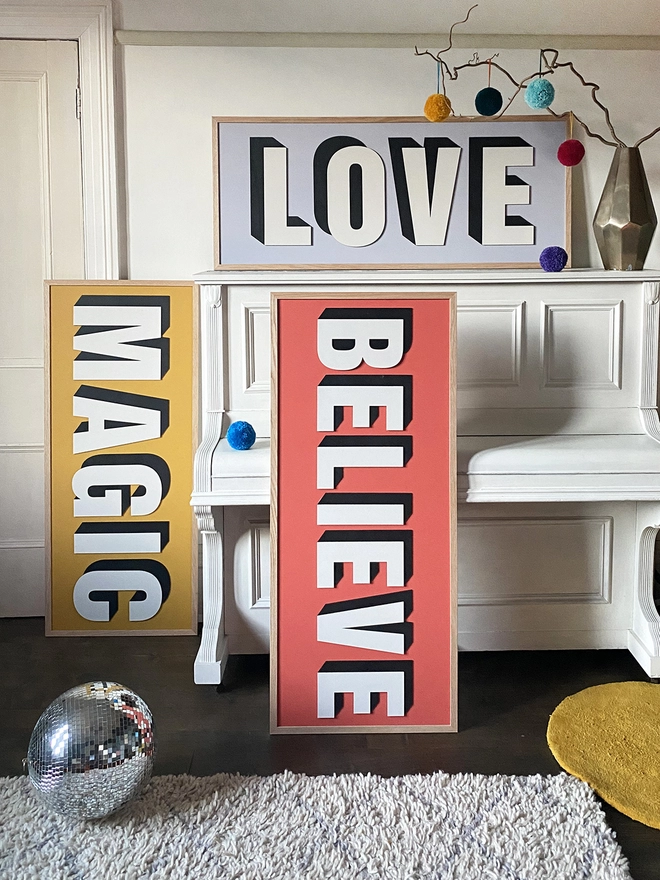 3 painted wooden sign leant up against a piano. one reading MAGIC with a yellow background, another reading BELIEVE on a red background & one reading LOVE on a lilac background.
