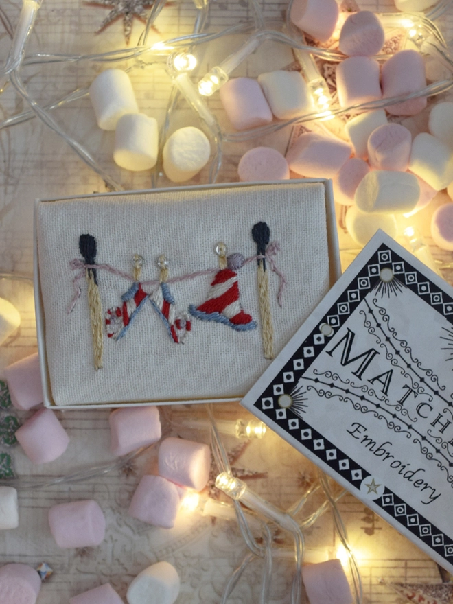 The elf laundry embroidery displayed in its matchbox, surrounded by mini marshmallows and fairy lights.