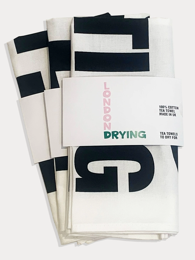A few white tea towels with black text laying flat on white background with London Drying 'belly band' around them to show packaging 