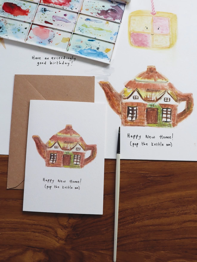 Desk Shot Of The Happy New Home Cottage Teapot Card Sitting Along Side The Original Hand Painted Watercolour Illustration, Paintbrush and Palette