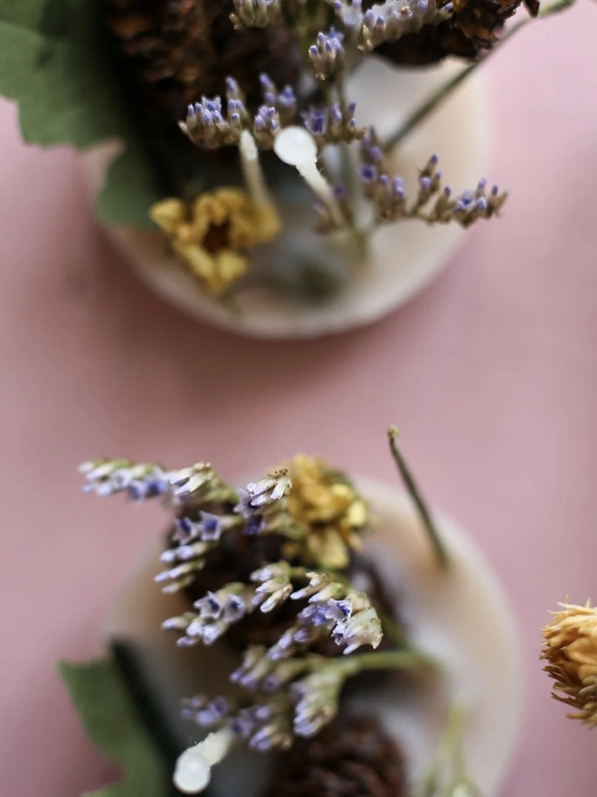 Recycled Wax Firelighters (Box Of 6). a close-up shot looking down on two of the recycled wax firelighters shows their pale wax bases topped with dried lavender, small yellow flowers and pine cones. the firelighters sit against a soft pink background, with the wicks visible poking up from the centre of each wax disc. a third firelighter is partially visible at the edge of the frame, its dried yellow bloom catching the light.