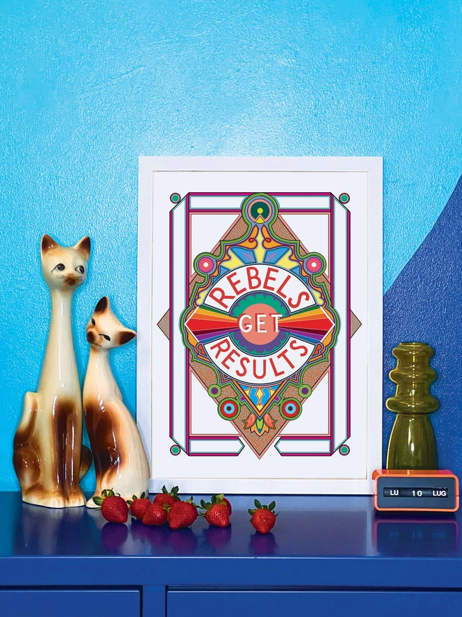 Rebels Get Results is written in red on a white background at the centre of this vibrant, abstract portrait illustration, with a white background and rainbows emitting from the centre, and multi-coloured detailing. The picture is in a white frame, against a turquoise and blue wall resting on a blue cabinet. Next to the picture are two cat ornaments, some ripe strawberries, a yellow glass vase and an orange Italian plastic calendar showing the date as ‘LU 10 LUG’.