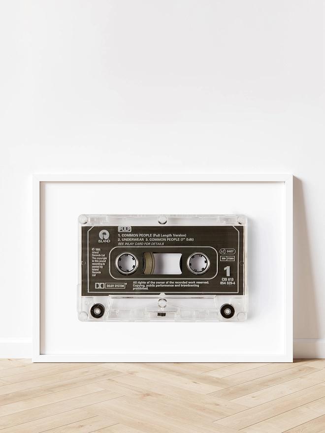 Pulp 'Common People' cassette tape print. framed print propped up against white wall and wooden floor.