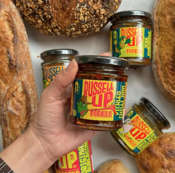 hand holding a jar of pickled sweet chili jam above a background of other jars of jam and loaves of bread