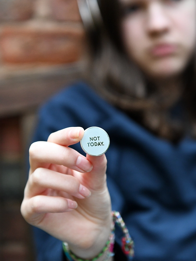 A teenage girl holds a round grey enamel pin badge in her hand that reads "Not Today".