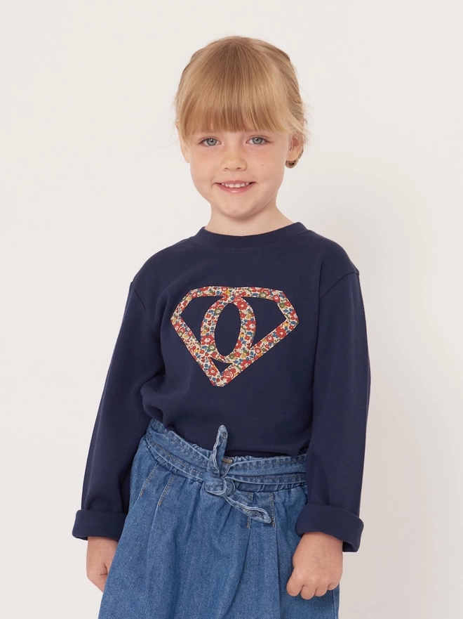 A girl wearing a navy long sleeve cotton t-shirt featuring the initial O inside a superhero motif. The motif is appliquéd in floral Liberty Print.
