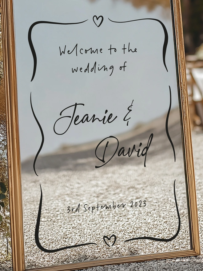 a gold framed mirror with the black writing of Welcome to the wedding of Jeanie and David on 3rd September 2026 with a heart at the top and one on the bottom and some squiggle lines around the words