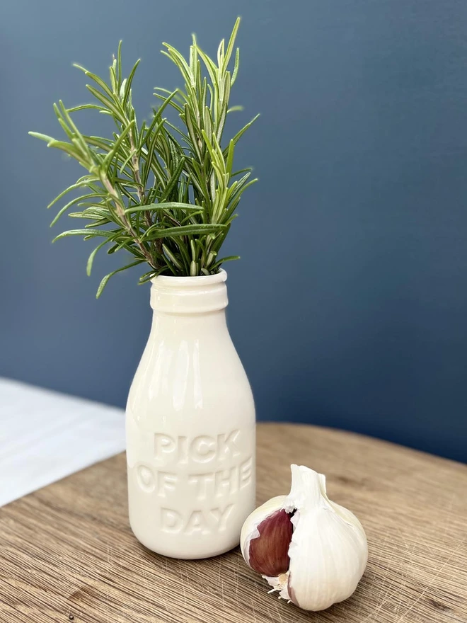 Small white ceramic bottle filled with rosemary sitting on a chopping board. Pick of the day is written on the side of bottle.