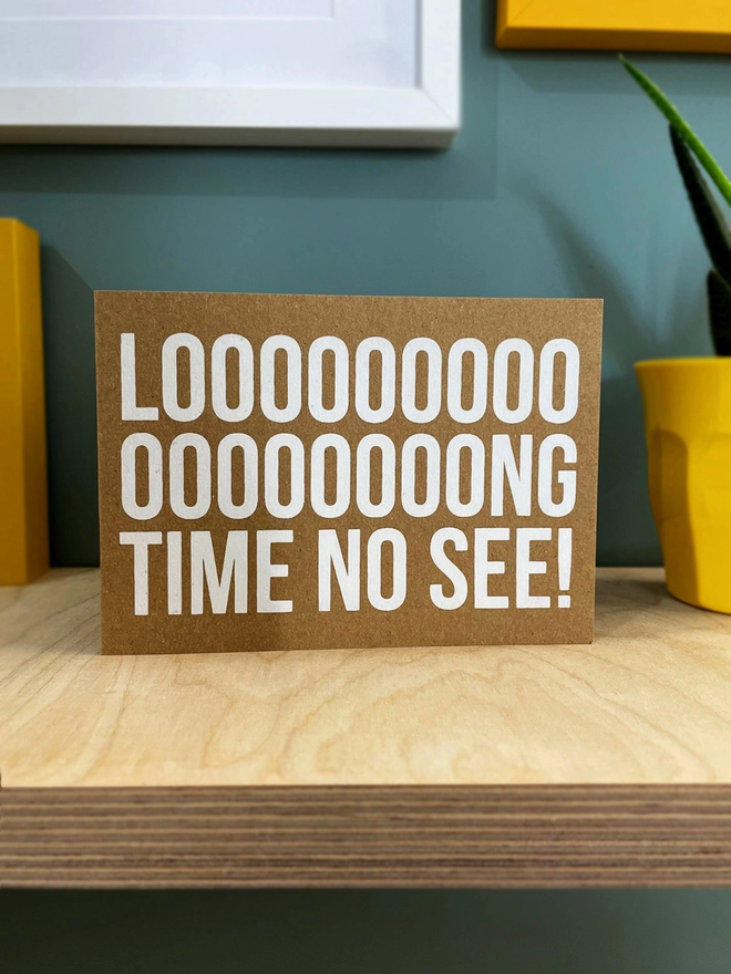 Looooooong time no see screenprinted across this card with multiple o’s in the Long, to emphasise the LONG. Printed in white ink on a brown kraft card stock in landscape format. Stood on a plywood shelf with hints of coloured frames around and a plant to one side. 