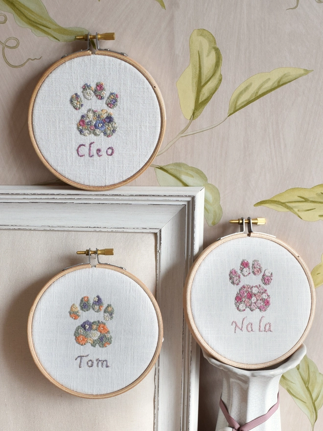 A display of 3 Cat Paw embroidery kits in their hoops, top to bottom are 1. Floral Meadow (Lavender Blues, Magenta & Buttermilk Yellow) 2. Pink Roses (5 shades of pink).  3. Sunshine Garden (Oranges, Yellows, Purples).