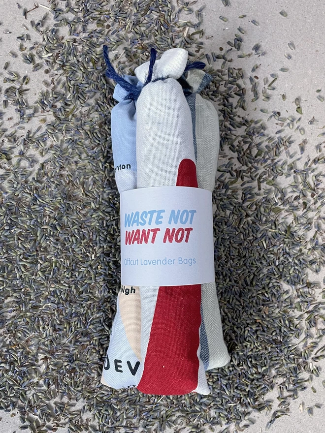 A bundle of three long style cotton lavender bags, tied at the tops, gathered with a branded cuff, sit on a pile of fresh lavender - it says Waste Not Want Not, Offcut Lavender Bags. The fabric has bits of red, blue and cream visible.