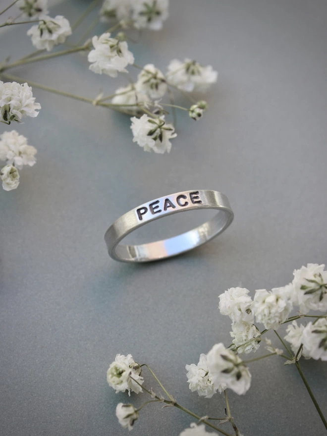 A sterling silver ring band with the word 'peace' stamped on it, on a green background with white flowers on either side.