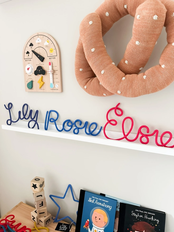 Three Personalised Kids Name Signs on a White Shelf in a Child's Playroom or Bedroom Wall with Wooden Alphabet blocks, toys and kid's books surrounding the knitted name signs. 