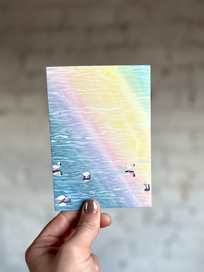 a greetings card featuring an illustration of flamingos in water with a rainbow reflection