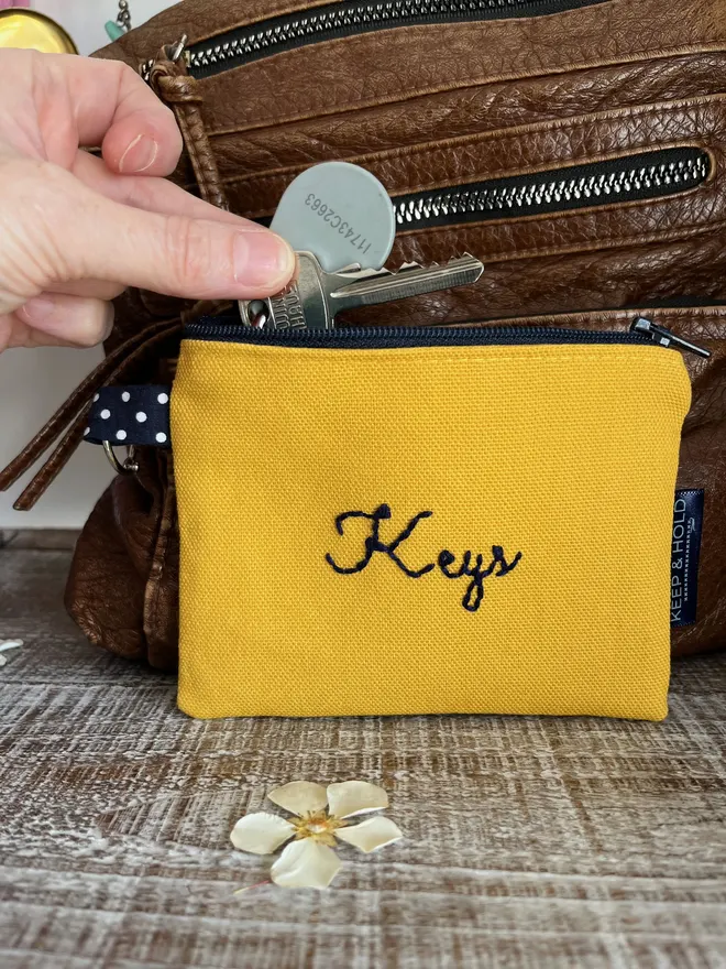 Small yellow coin purse with key ring & 'Keys' embroidered in navy thread. A hand is placing keys inside the open zipper.