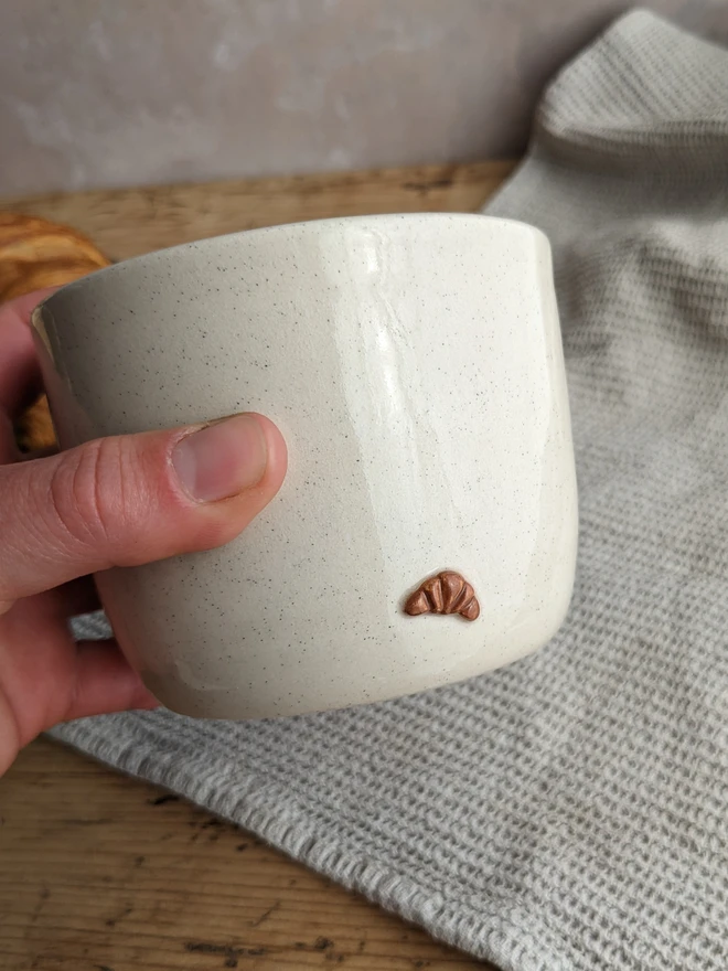 A small white plant pot with croissant design, holding a green leafy plant on a wooden surface.
