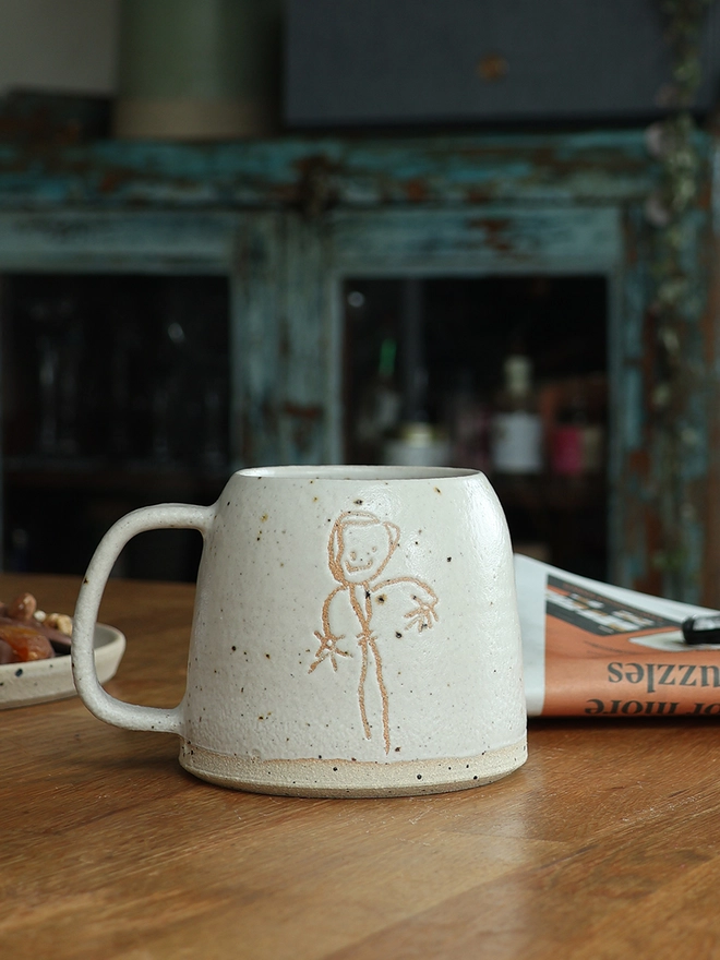 Childrens Drawing Handmade Ceramic Mug sat on a wooden table with a plate of biscuits to its right and a newspaper to its left