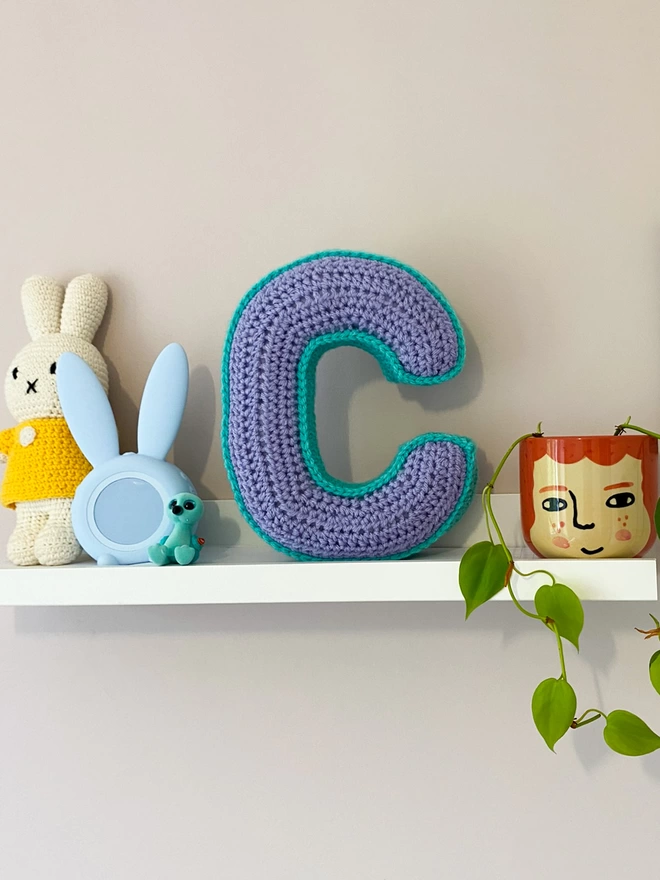 Crocheted C cushion in Lilac and Teal on childs shelf