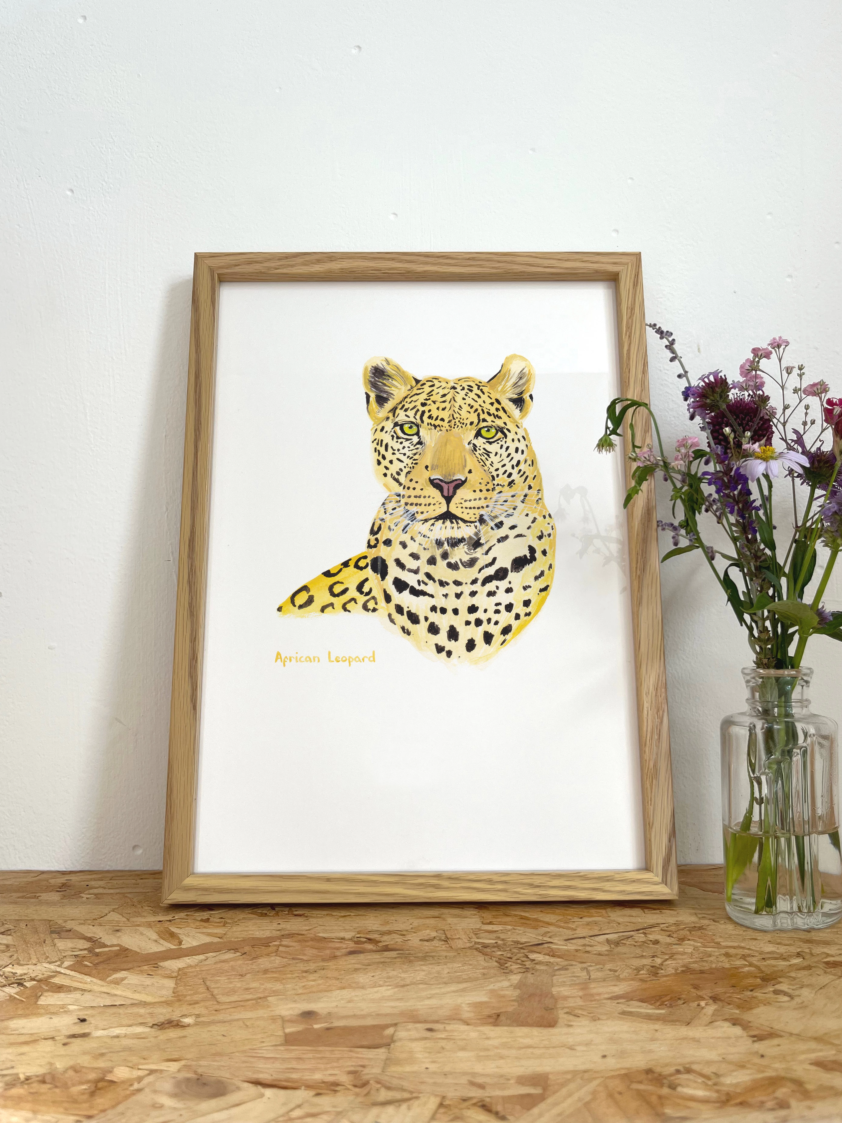 A print of an illustrated head and chest of an African Leopard in a frame next to some flowers