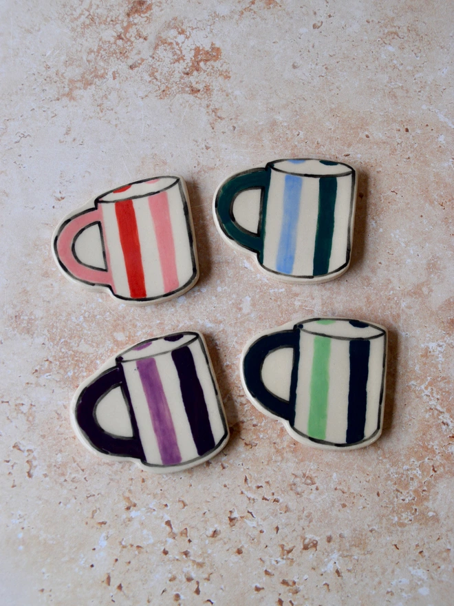 Handmade ceramic mug-shaped magnets with bold stripe patterns in red, pink, blue, green and purple, displayed on a neutral stone background.