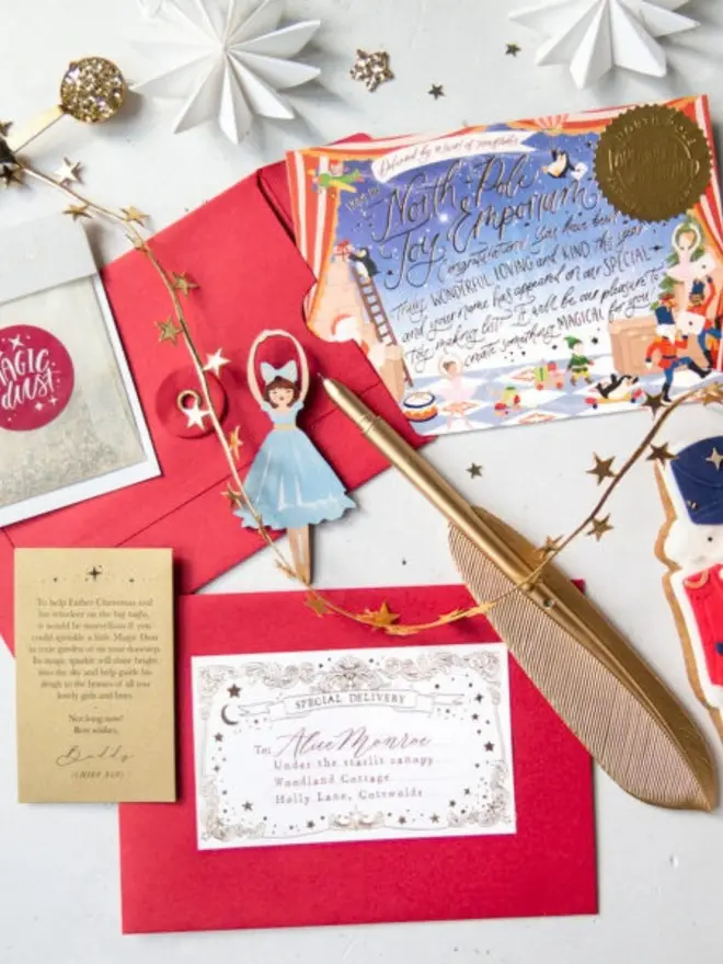 A letter from Father Christmas kit with red envelopes, a personalised "Special Delivery" label, a gold pearlescent Magic Dust instruction card and the illustrated North Pole notecard, arranged with a gold feather pen, a ballerina decoration and a gold star garland.