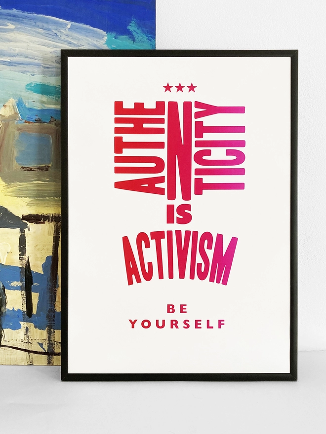 Framed multicoloured typographic print of the quote “Authenticity Is Activism”  The print rests against a blue and yellow abstract painting.