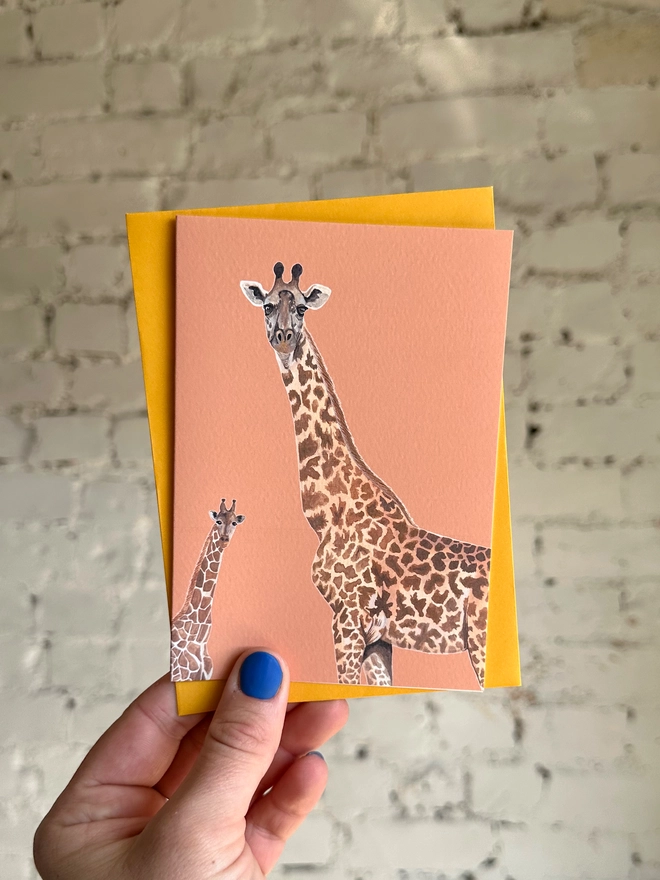an orange greetings card featuring illustrations of two giraffes with a deep yellow envelope