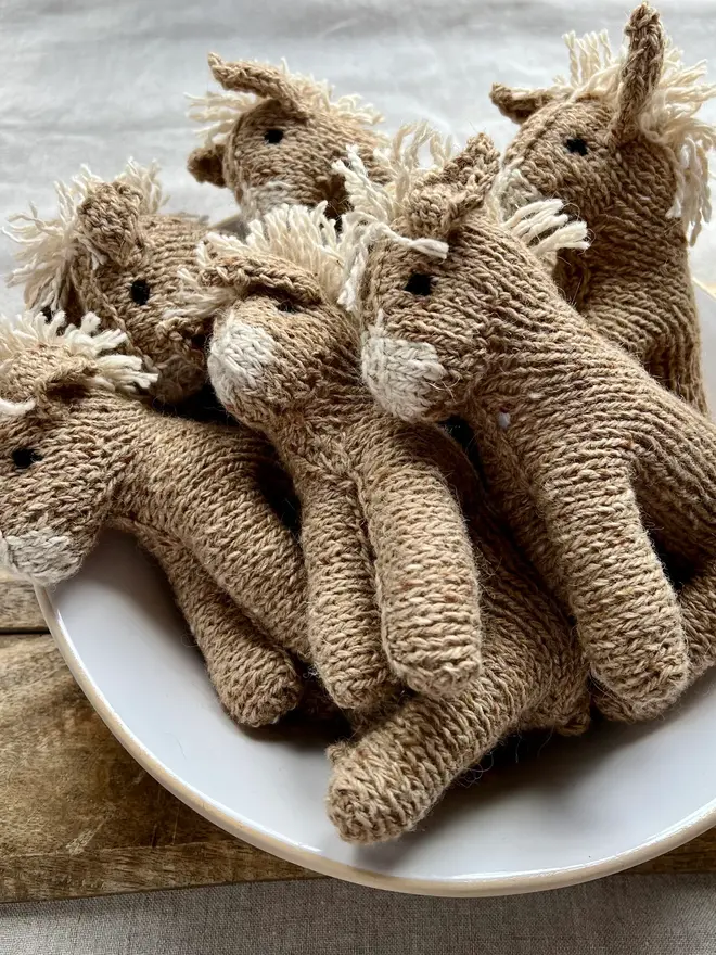 Hand Knitted Natural Dyed Wool And Alpaca Yarn Donkey Gift, a group of hand knitted donkeys sitting in a white bowl on a wooden chopping board on a table cloth. They are accompanied by some hey and some knitting accessories. 