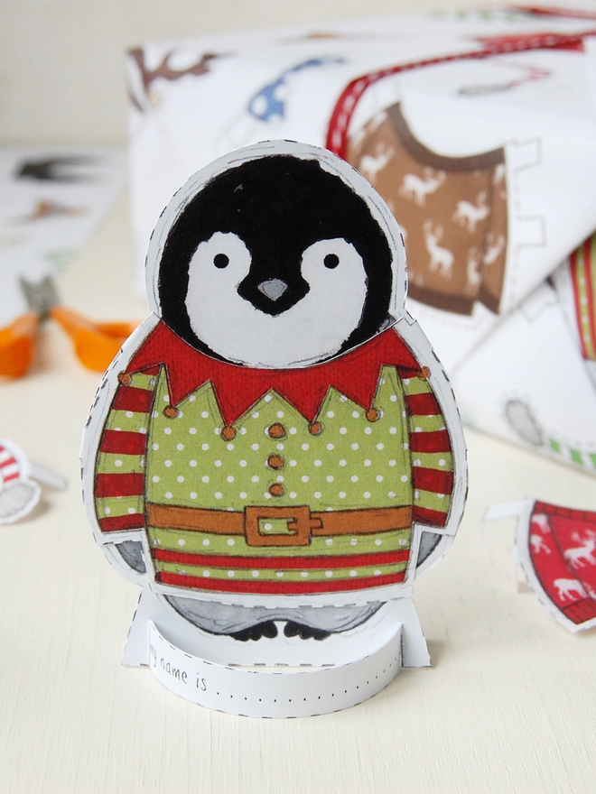 A gift wrapped in 'dress up a penguin' paper doll Christmas wrapping paper is on a white wooden table.