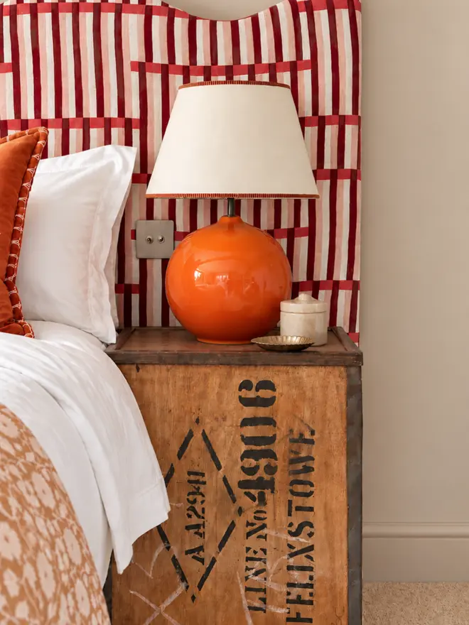 Bright, colourful bedroom interior featuring a glossy orange ceramic table lamp with a cream empire lampshade, styled on a vintage wooden trunk bedside table. A red and pink striped upholstered headboard, crisp white bedding and rust-coloured cushions create a warm, eclectic bedroom scheme.