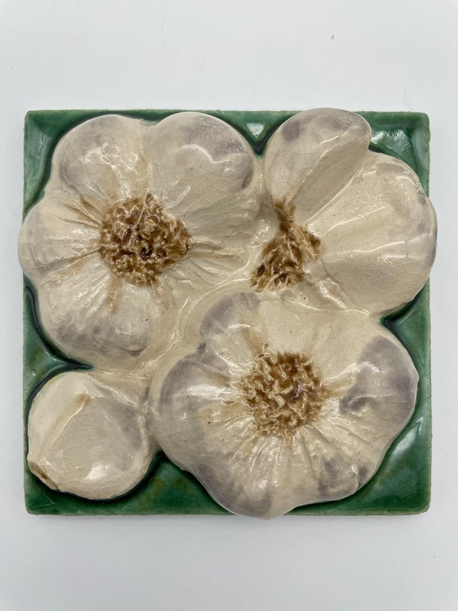 Handmade ceramic tile taken from a plaster cast of real bulbs of garlic, bottom view, plus a single clove. Very realistic, three-dimensional, with lush coloured glazes.