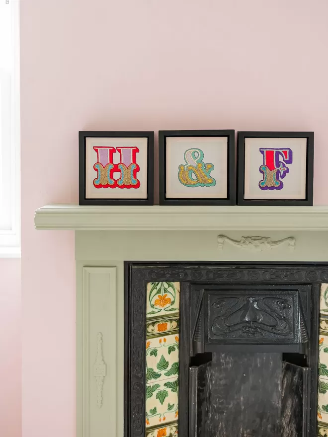 Hand embroidered initials and ampersand in frames, against a pink wall, and on a painted original fireplace